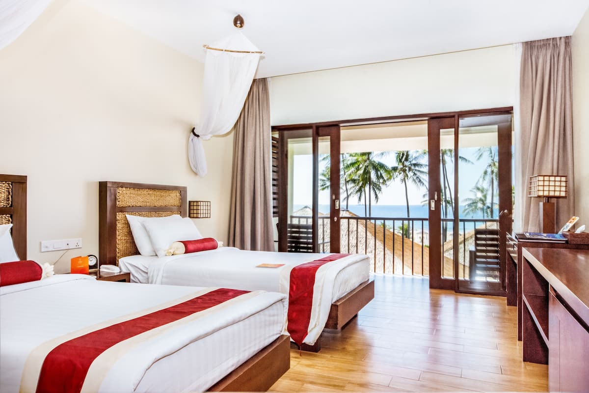 Une chambre d’hôtel avec deux lits, un balcon donnant sur des palmiers et l’océan, et un bureau en bois avec une lampe.
