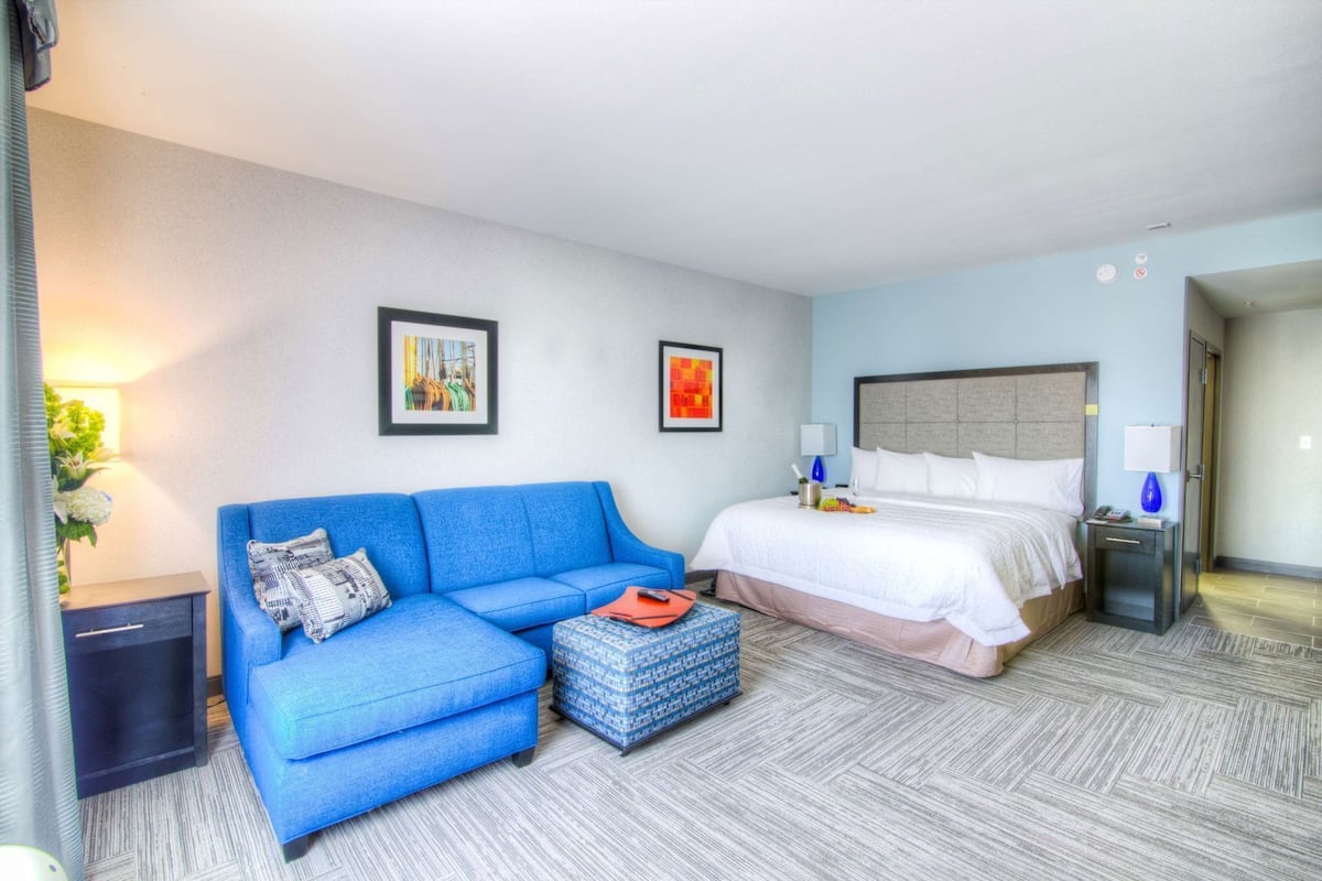 Habitación de hotel con sofá azul, cama, mesita de noche, lámpara y dos cuadros en la pared.