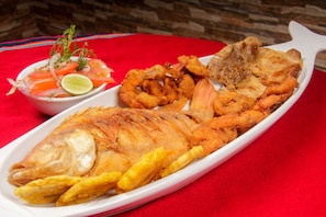 Un plato de pescado frito acompañado de aros de cebolla, plátanos fritos y un pequeño bol de ensalada.