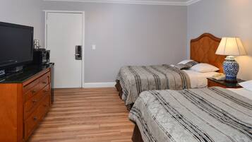 A hotel room with a wooden floor, a television on a dresser, two beds with striped bedding, a nightstand with a lamp, and a door.
