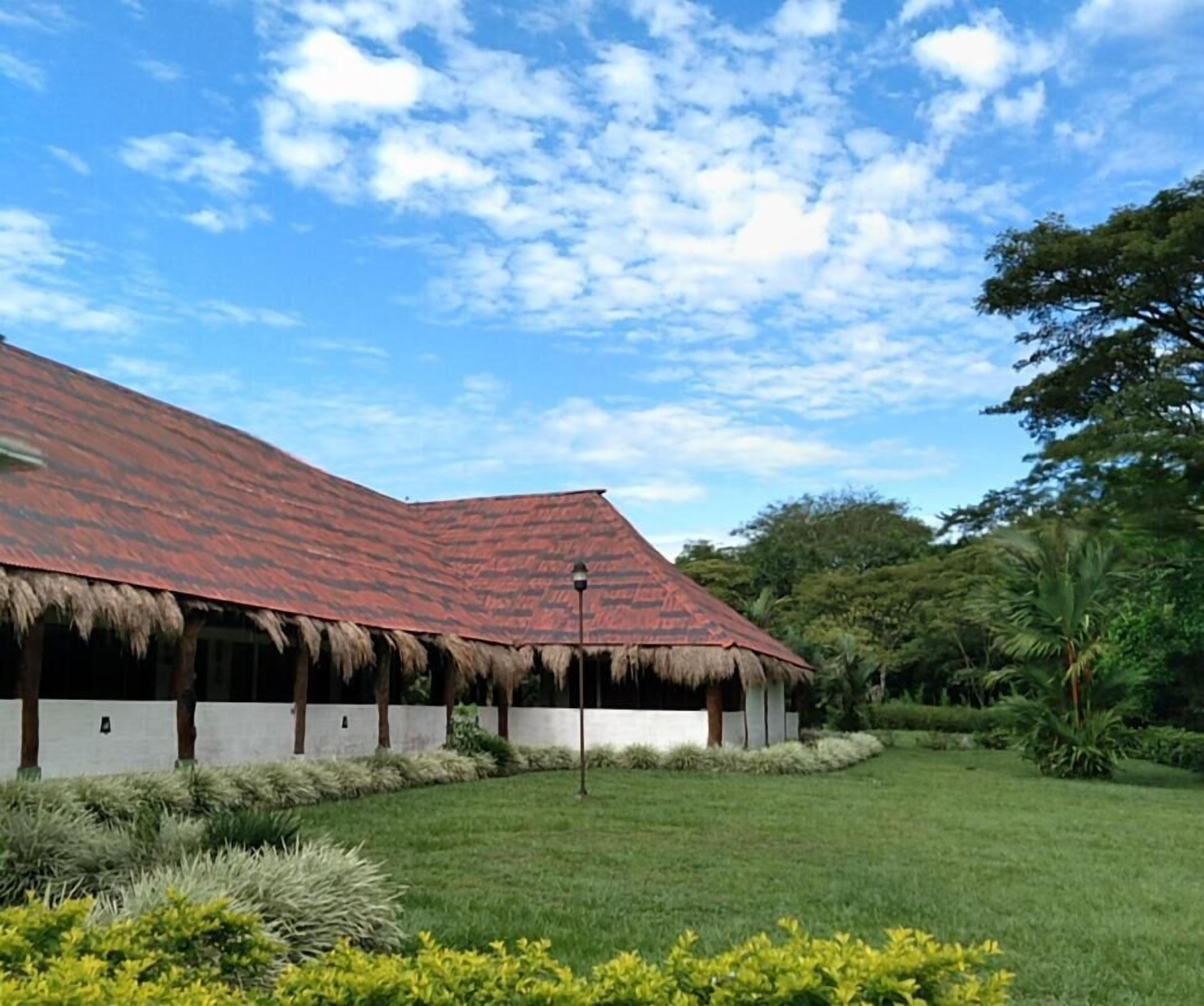 Un edificio con tejado rojo y aleros de paja, rodeado de césped cuidado y vegetación tropical.