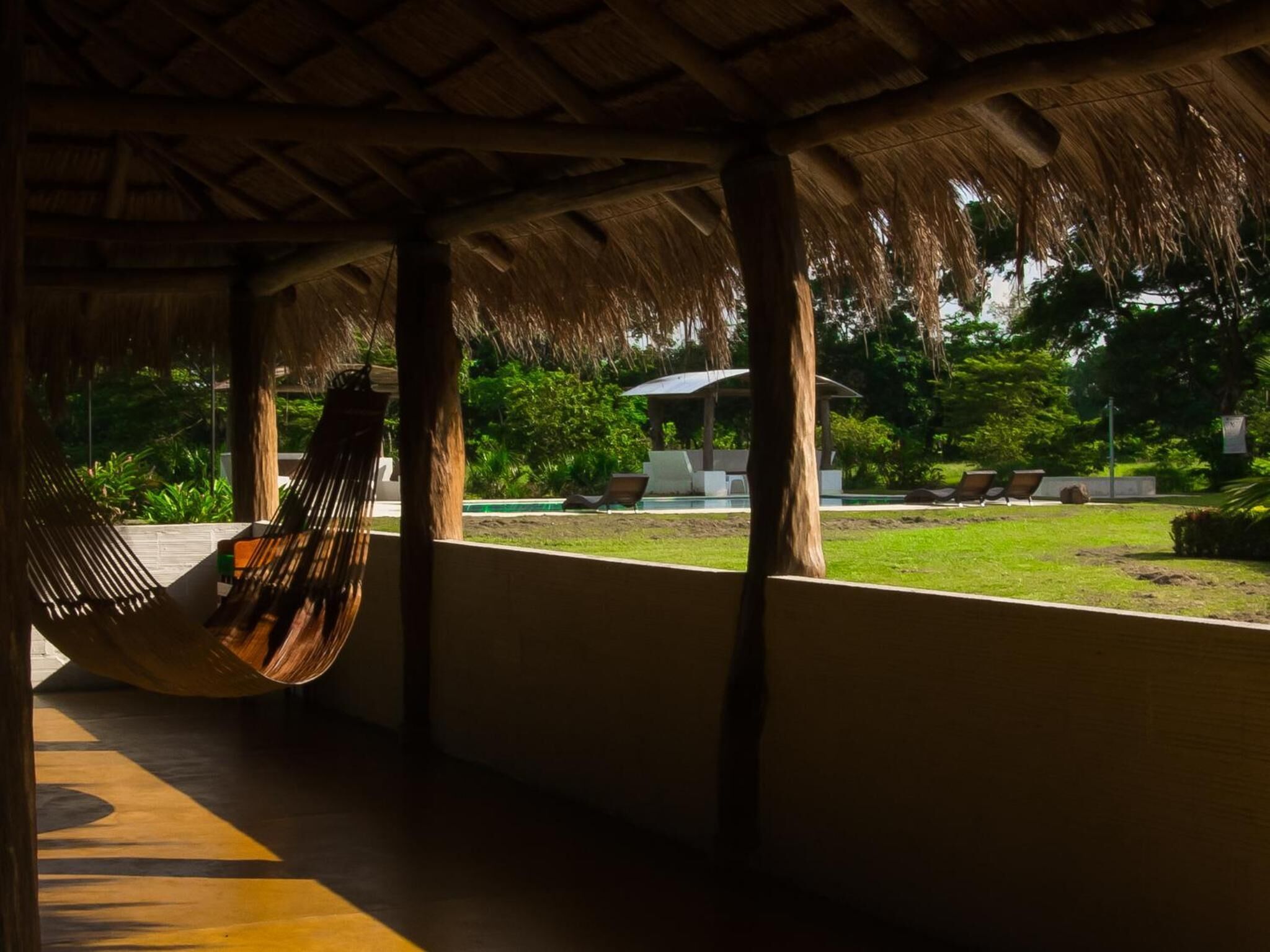 Una hamaca bajo un techo de paja, una piscina y un jardín con sillones de descanso.