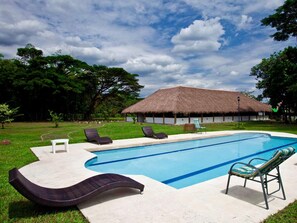 Una piscina con sillones de descanso, un edificio de techo de paja y abundante vegetación.