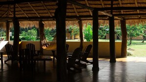 An outdoor covered seating area with tables and chairs, a hammock, and a view of a garden with trees and a building in the background. - Cinaruco Caney (Villavicencio)