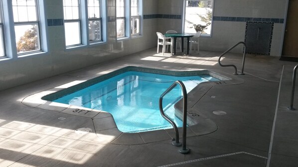 Una piscina rectangular interior con una marca de profundidad de 90 centímetros, rodeada de suelo y paredes con azulejos, y una mesa con sillas al fondo.