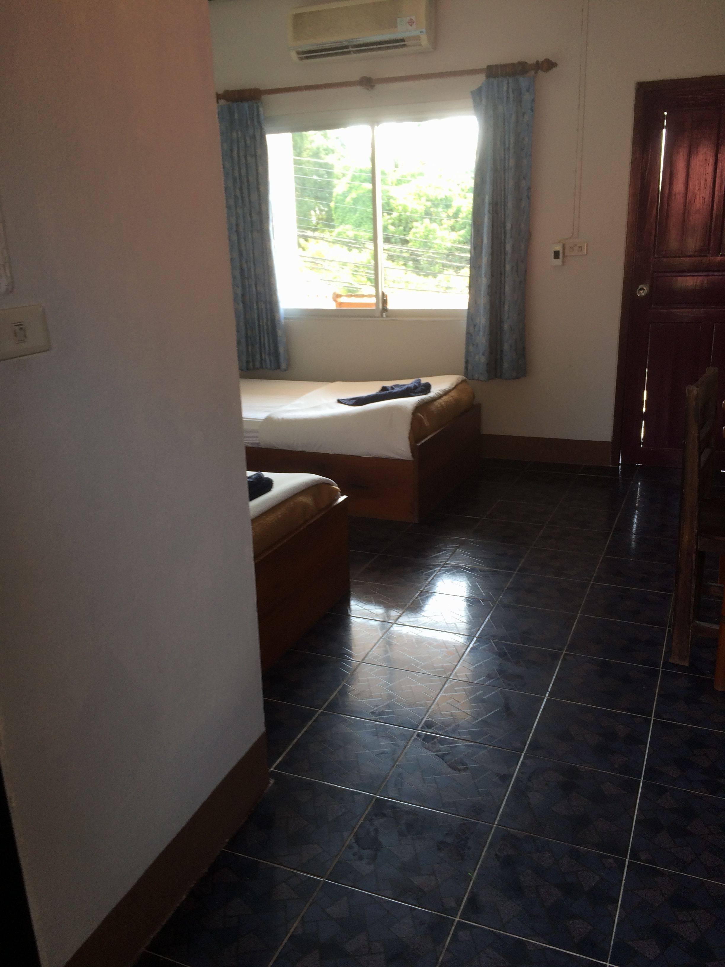 A room with two beds, a window with curtains, a tiled floor, and a wooden door.