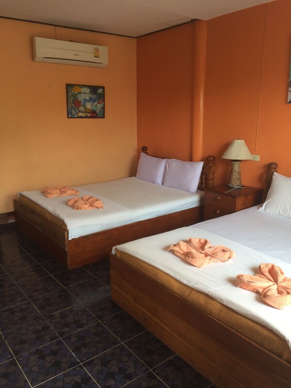 Habitación de hotel con dos camas individuales, cada una con una almohada y una toalla. Hay una mesita de noche con una lámpara y una pequeña mesa. La habitación tiene paredes anaranjadas y suelo de baldosas.