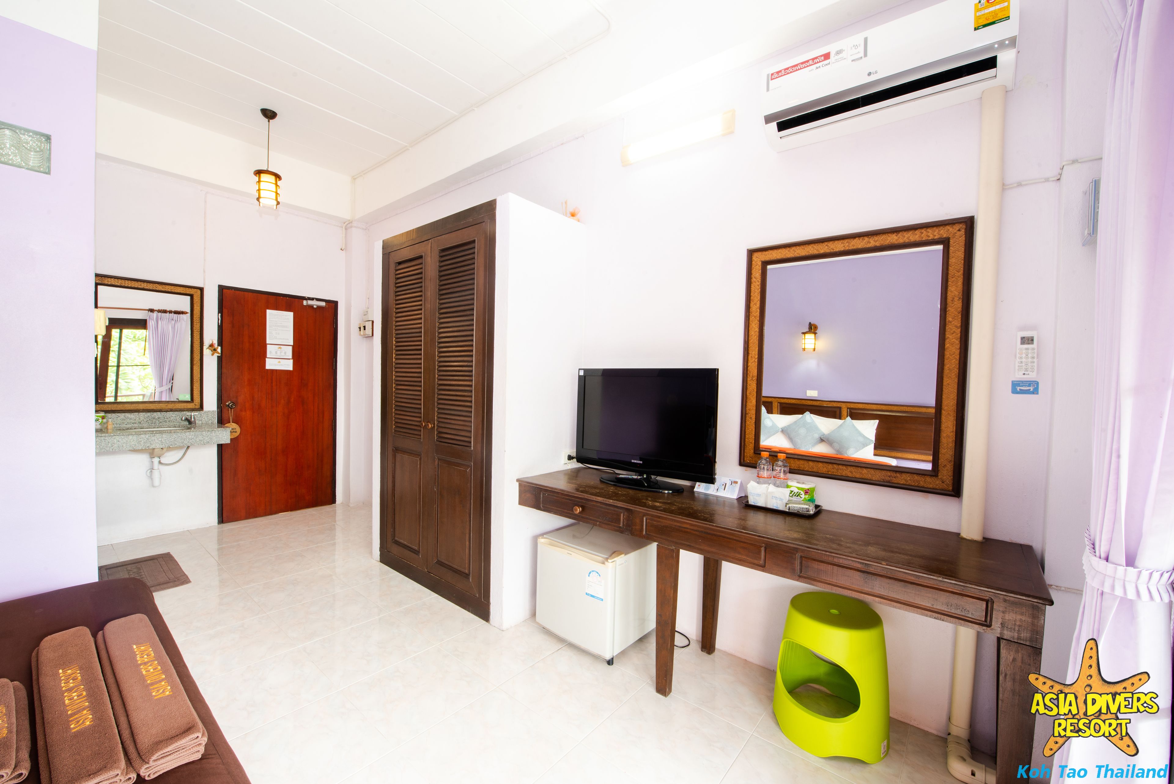 A hotel room with a wooden desk, a television, a small refrigerator, a green stool, a mirror, and a window with curtains.