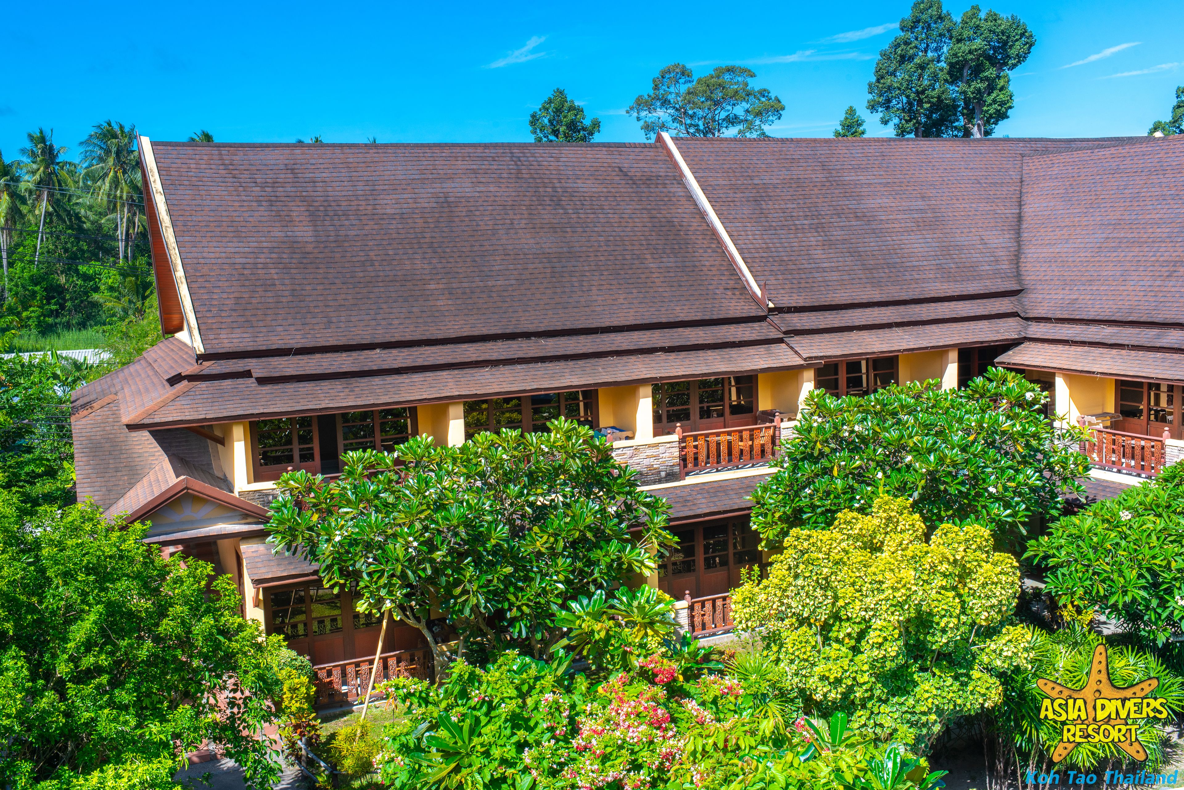 A two-story building with a sloped roof, multiple balconies, and lush greenery surrounding it.