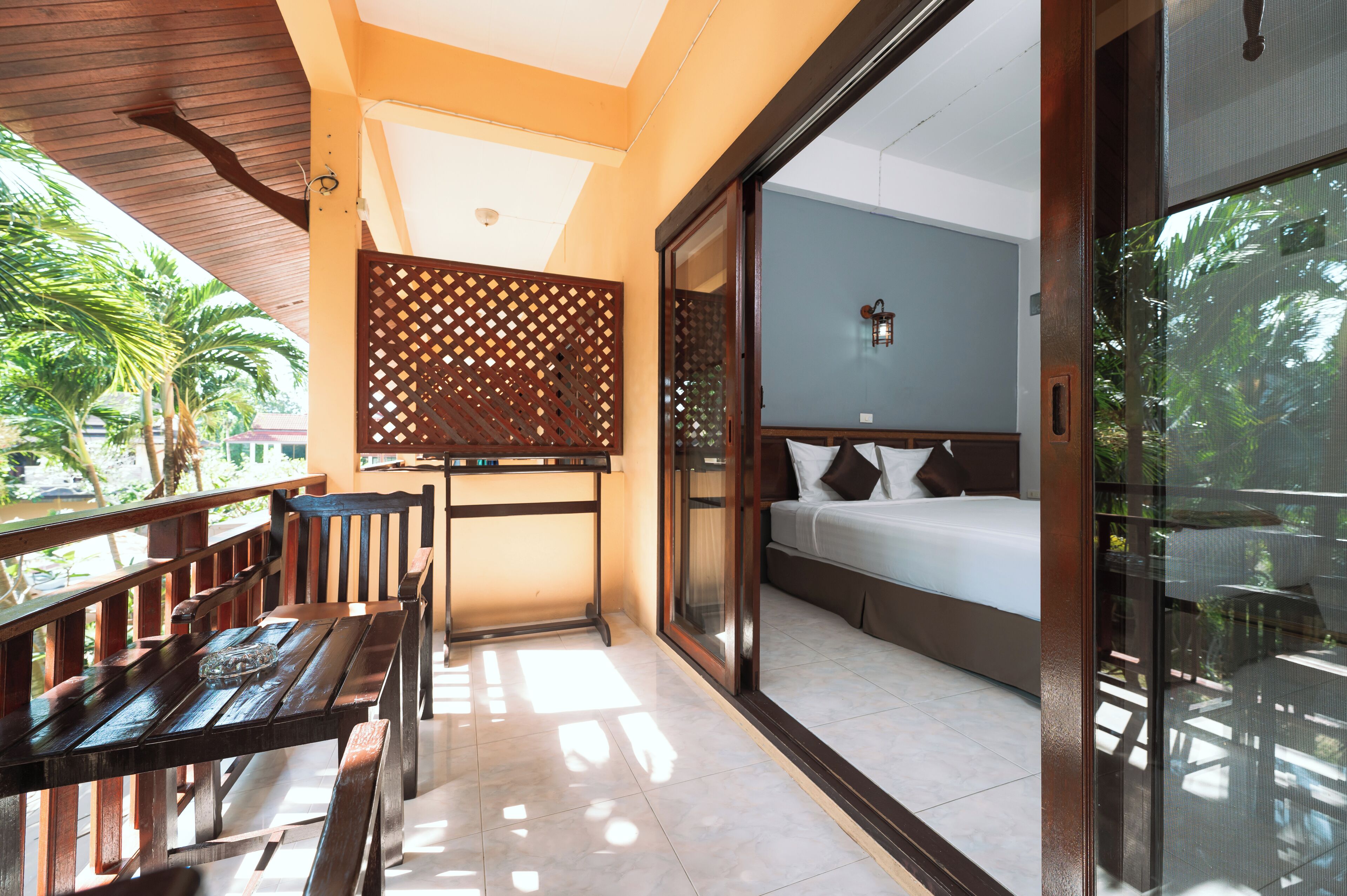 A balcony with a wooden table and chairs, a bed with a view of palm trees, and a sliding glass door.