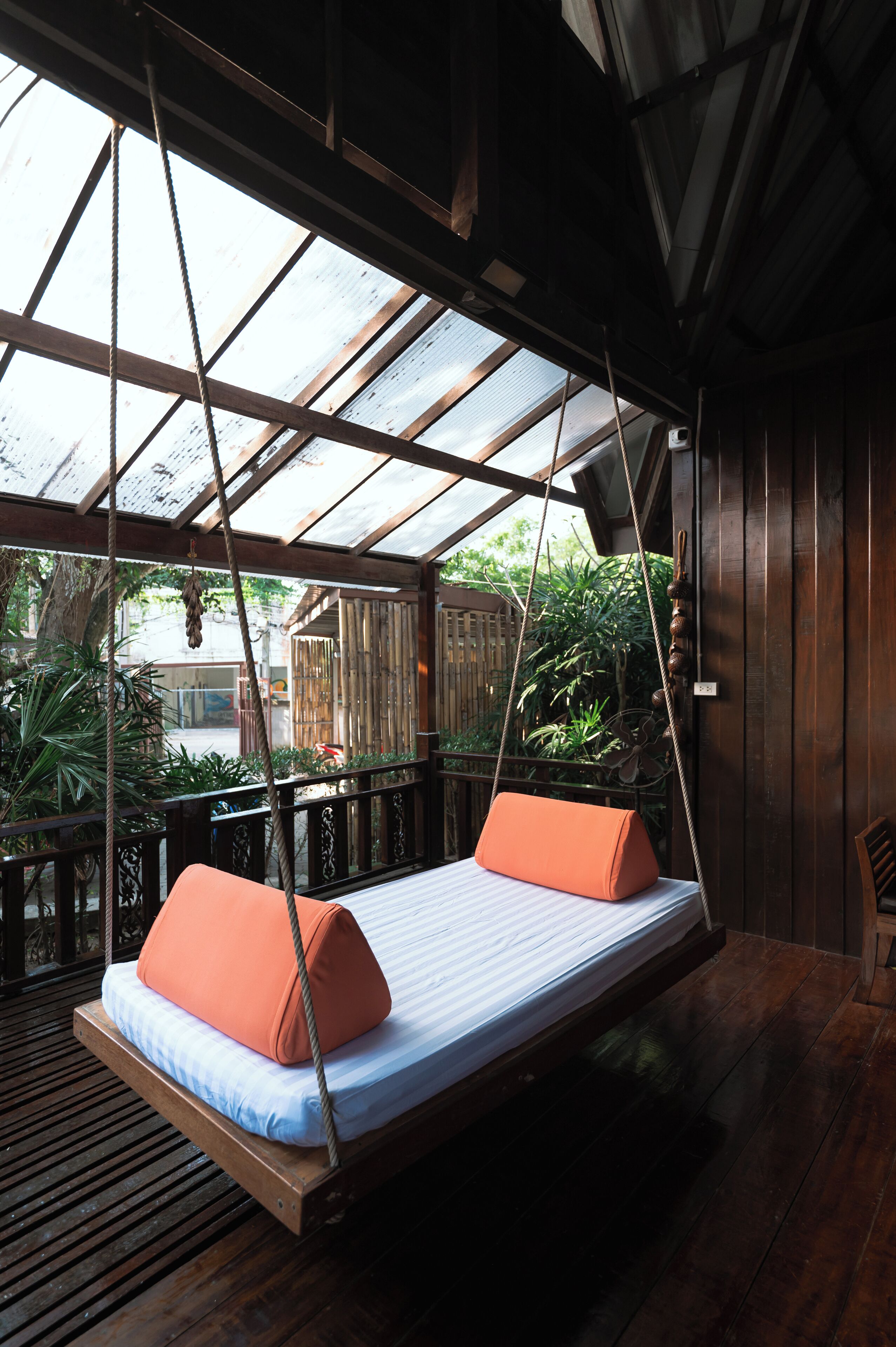 A wooden swing bed with orange cushions, a wooden floor, and a view of greenery outside.