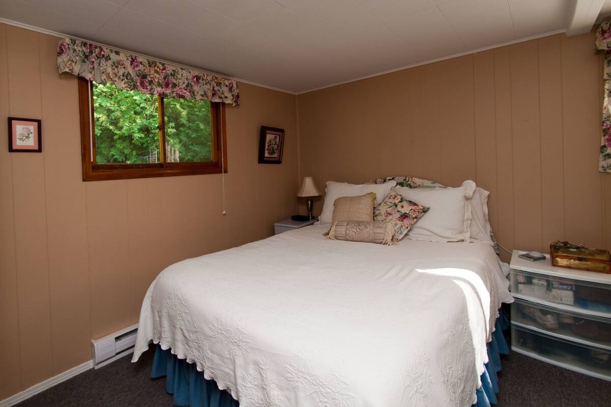 Una cama bien hecha con cubrecama blanco y almohadas florales, una mesita de noche con lámpara y una ventana con cortina de encaje floral.