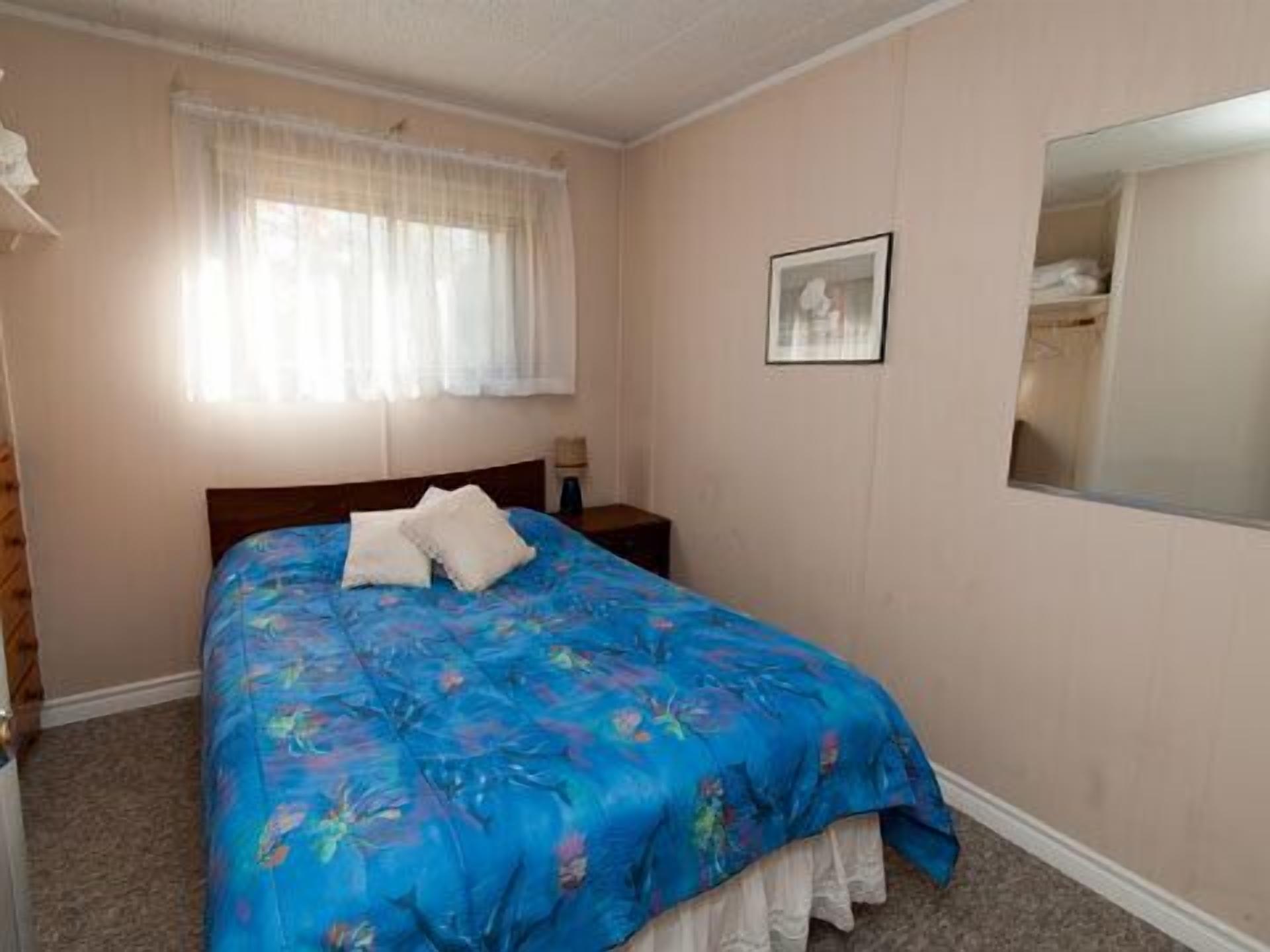 Un dormitorio con cama, mesita de noche, ventana con cortinas e imagen enmarcada en la pared.