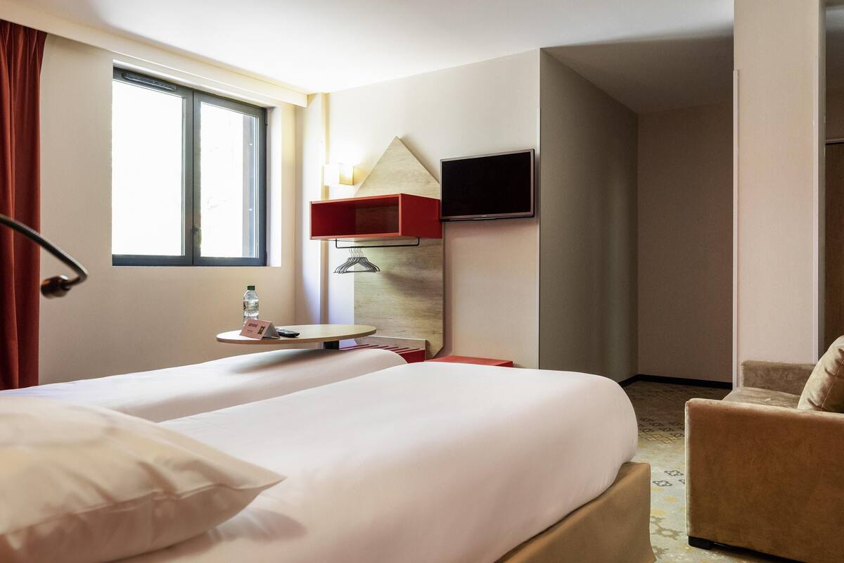 A hotel room with a bed, a chair, a TV, a window, and a table with a bottle and a sign.