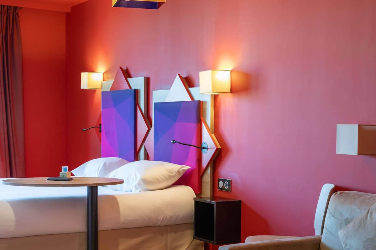 A hotel room with a bed, a bedside table, a chair, and a wall-mounted TV.