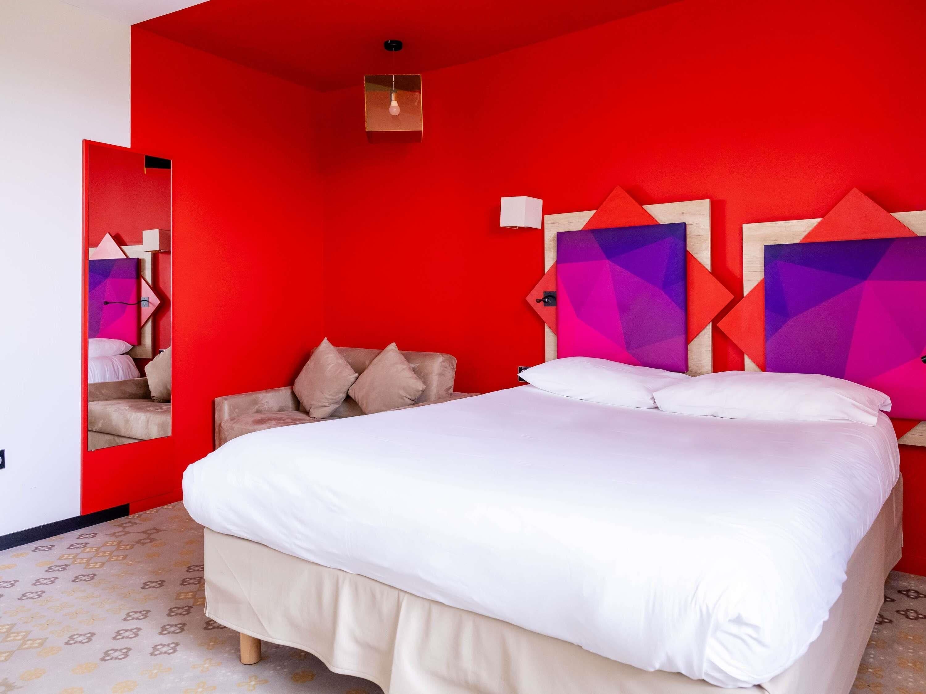 Un dormitorio moderno con una pared roja, una cama grande con ropa de cama blanca y cabeceros con motivos geométricos.