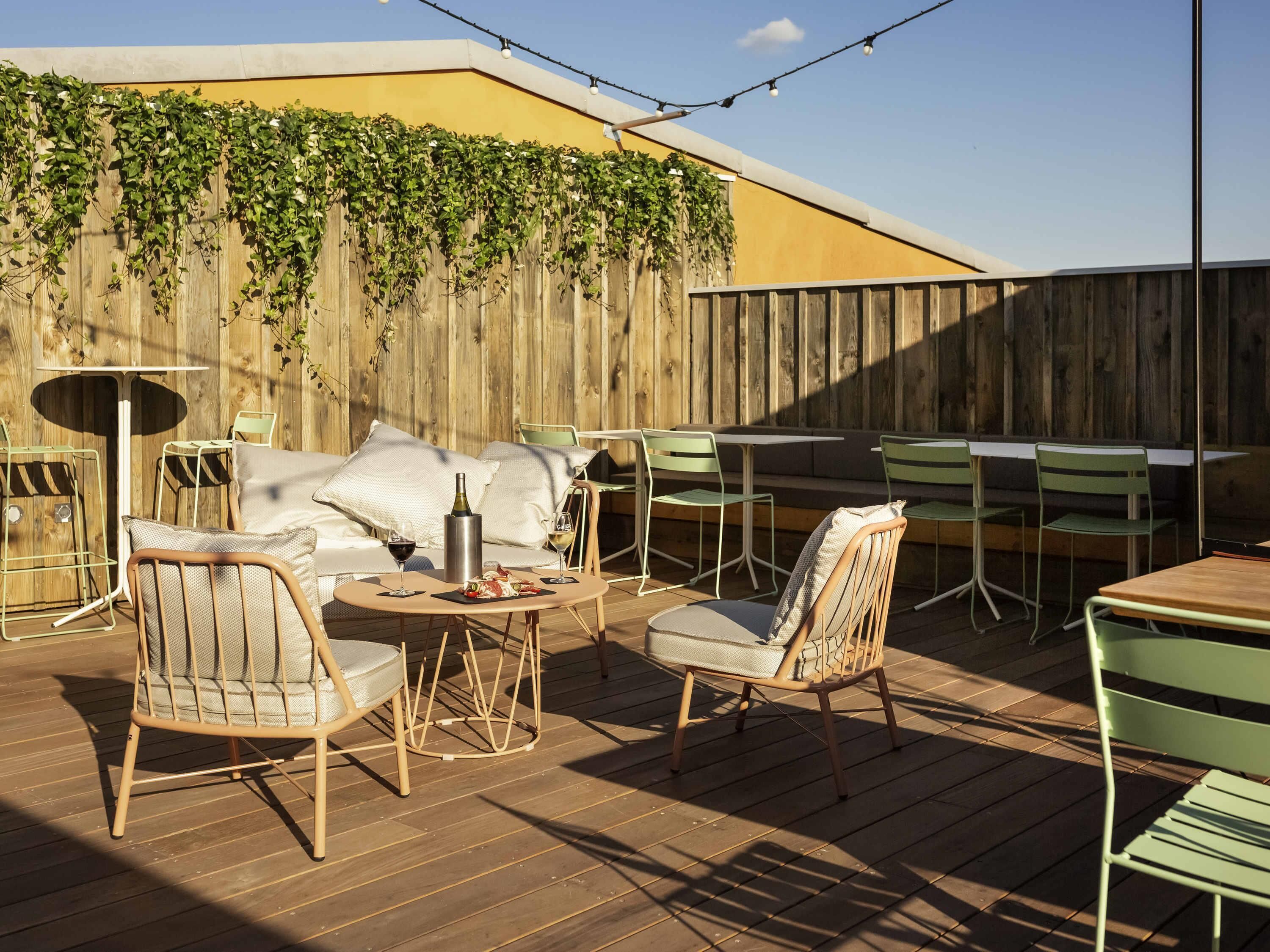 Un patio al aire libre con muebles de madera, una mesa con una botella y vasos, y una valla de madera cubierta de plantas trepadoras.
