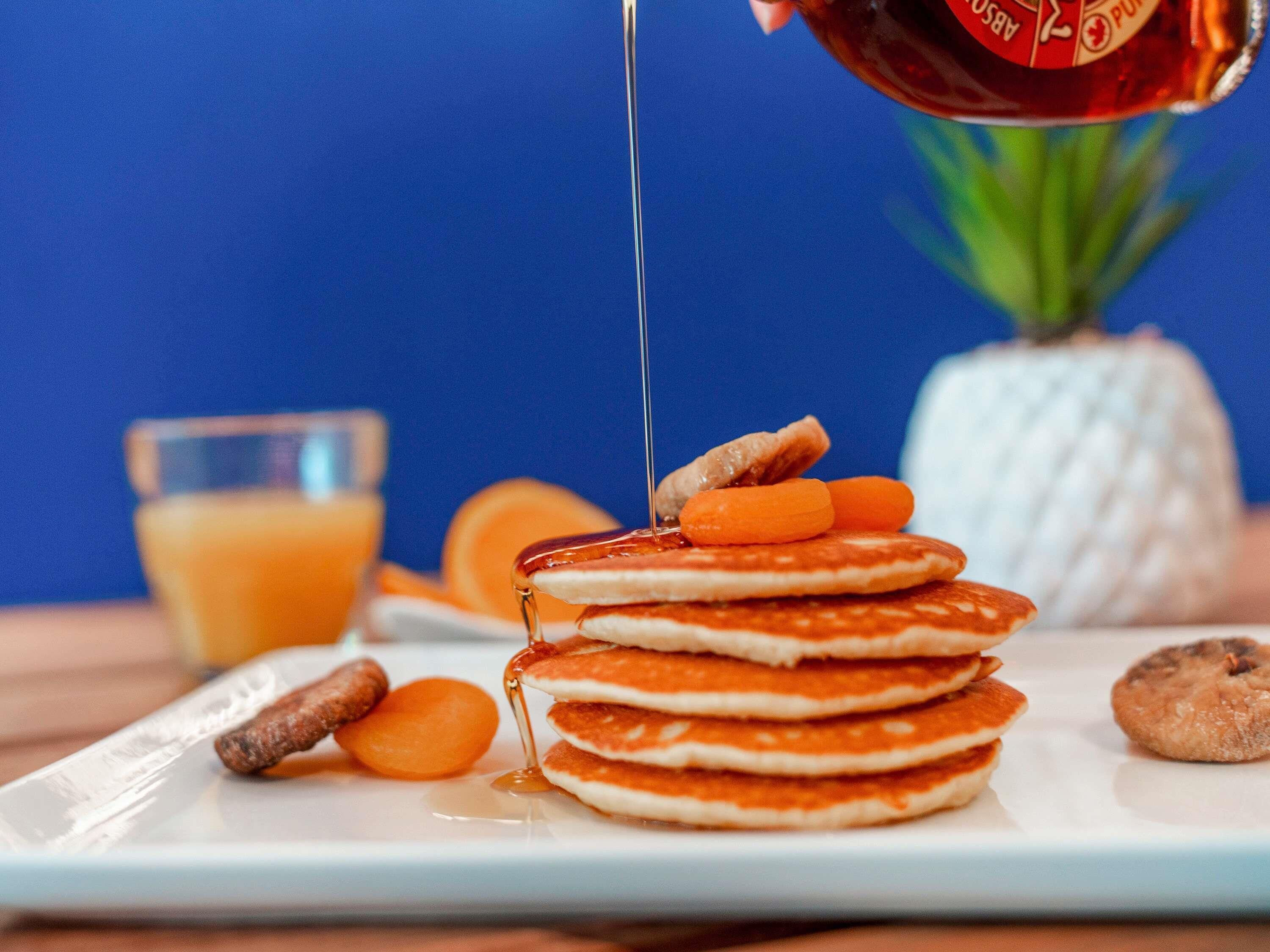 A stack of pancakes with syrup being poured over them, accompanied by a pineapple, orange slices, and a glass of orange juice.
