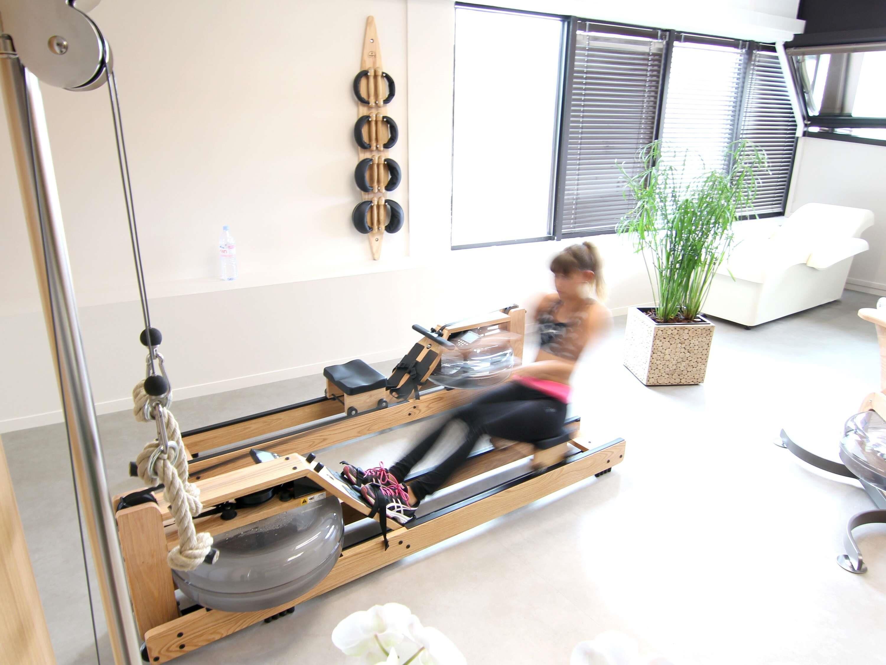 A Pilates reformer machine in a room with a large window, a white couch, and a potted plant.