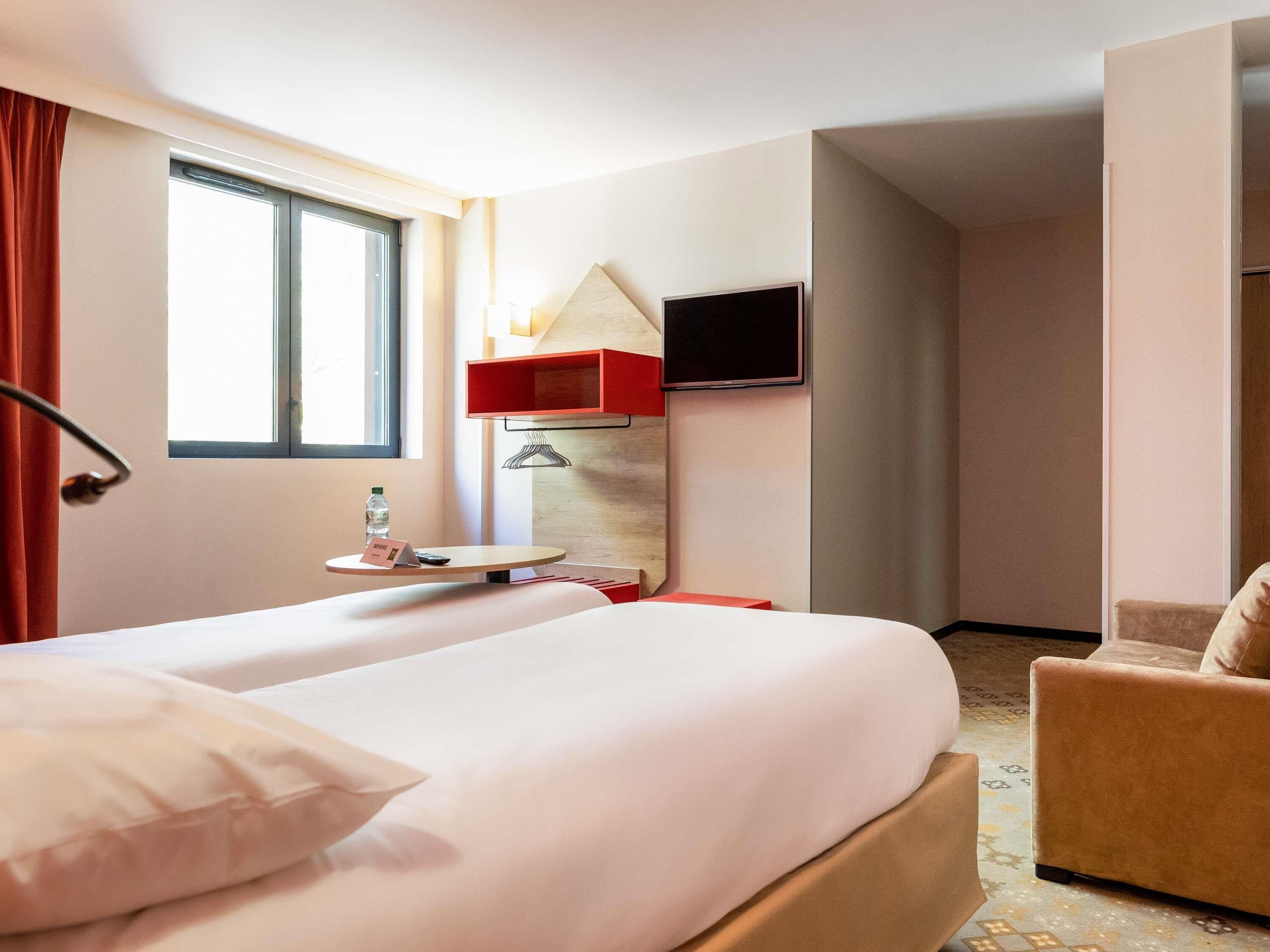 A hotel room with a bed, a chair, a TV mounted on the wall, a window with curtains, and a small table with a bottle and a sign.