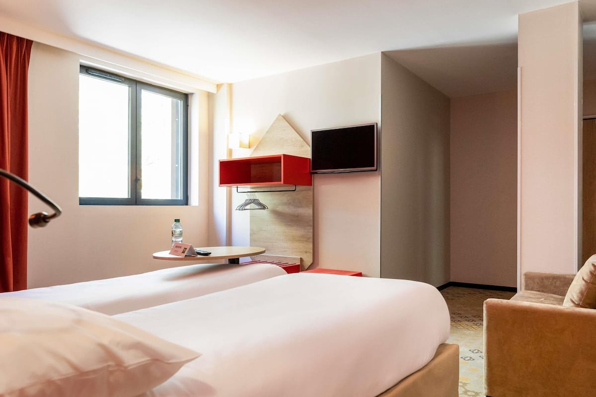 A hotel room with a bed, a chair, a TV mounted on the wall, and a window with curtains.