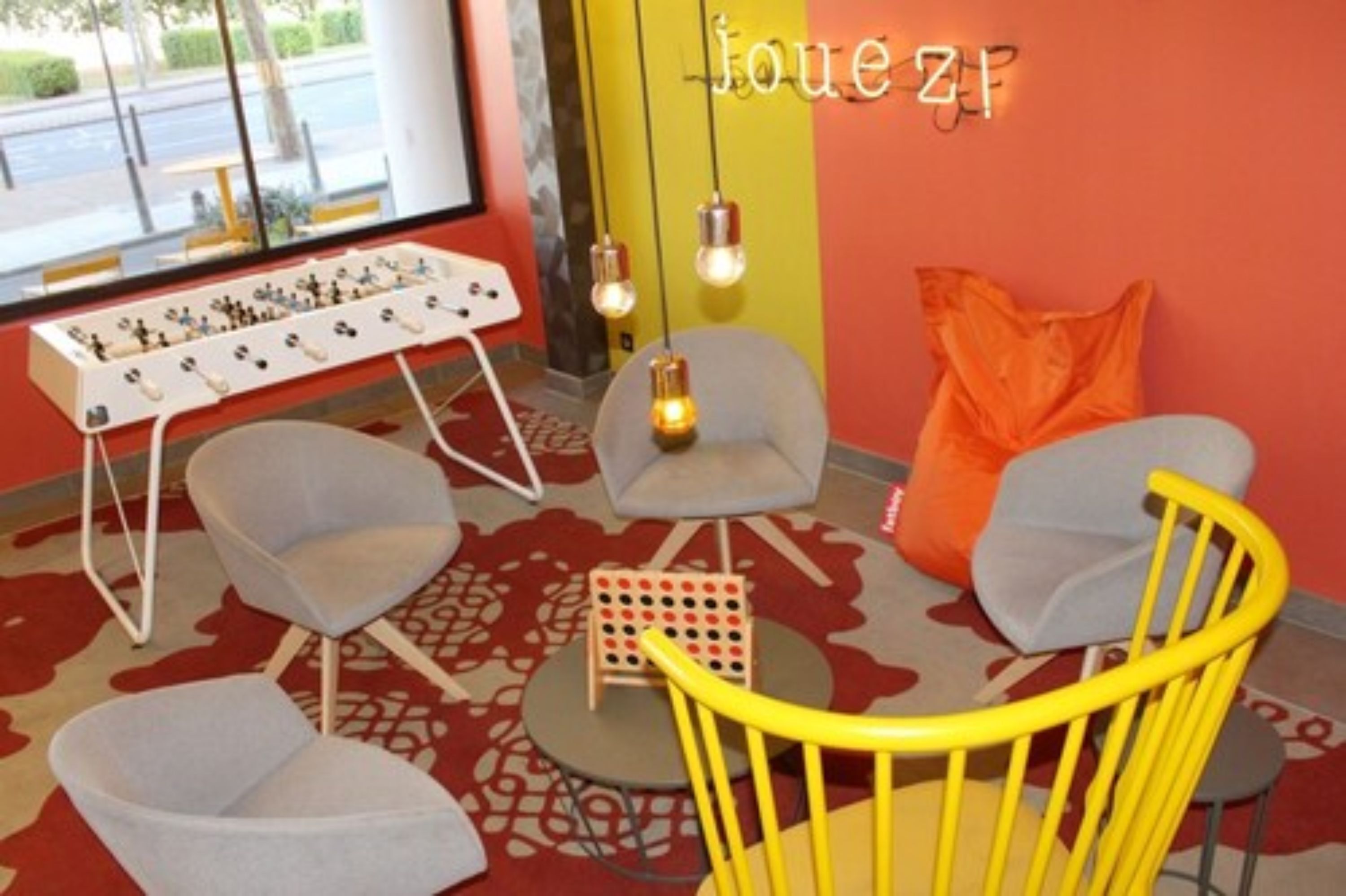 A room with a red wall, a yellow chair, a white table with a board game, and a window with a view of the street.