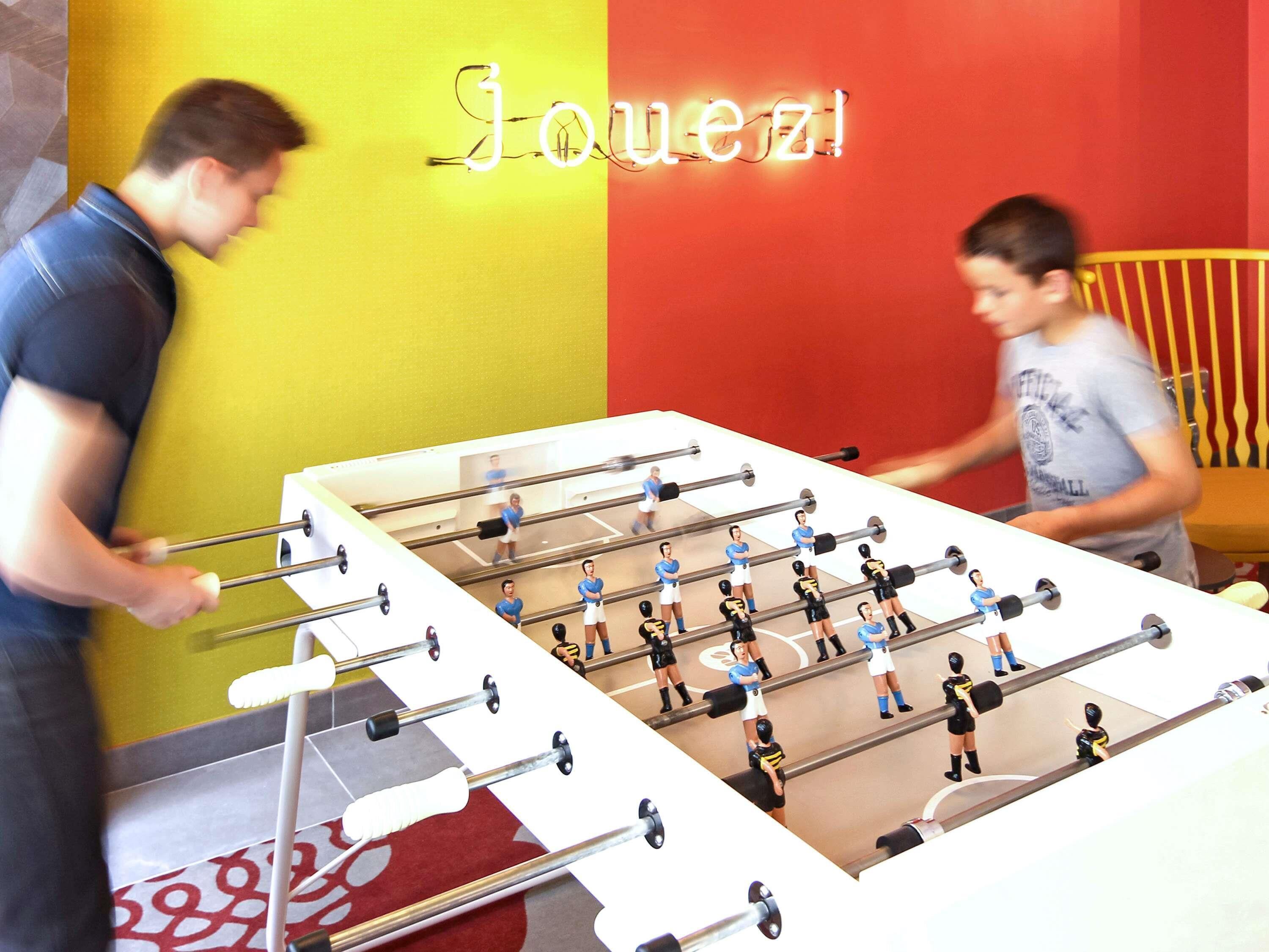 A foosball table in a room with a yellow and red wall, and a neon sign that reads 'Jouezi'.