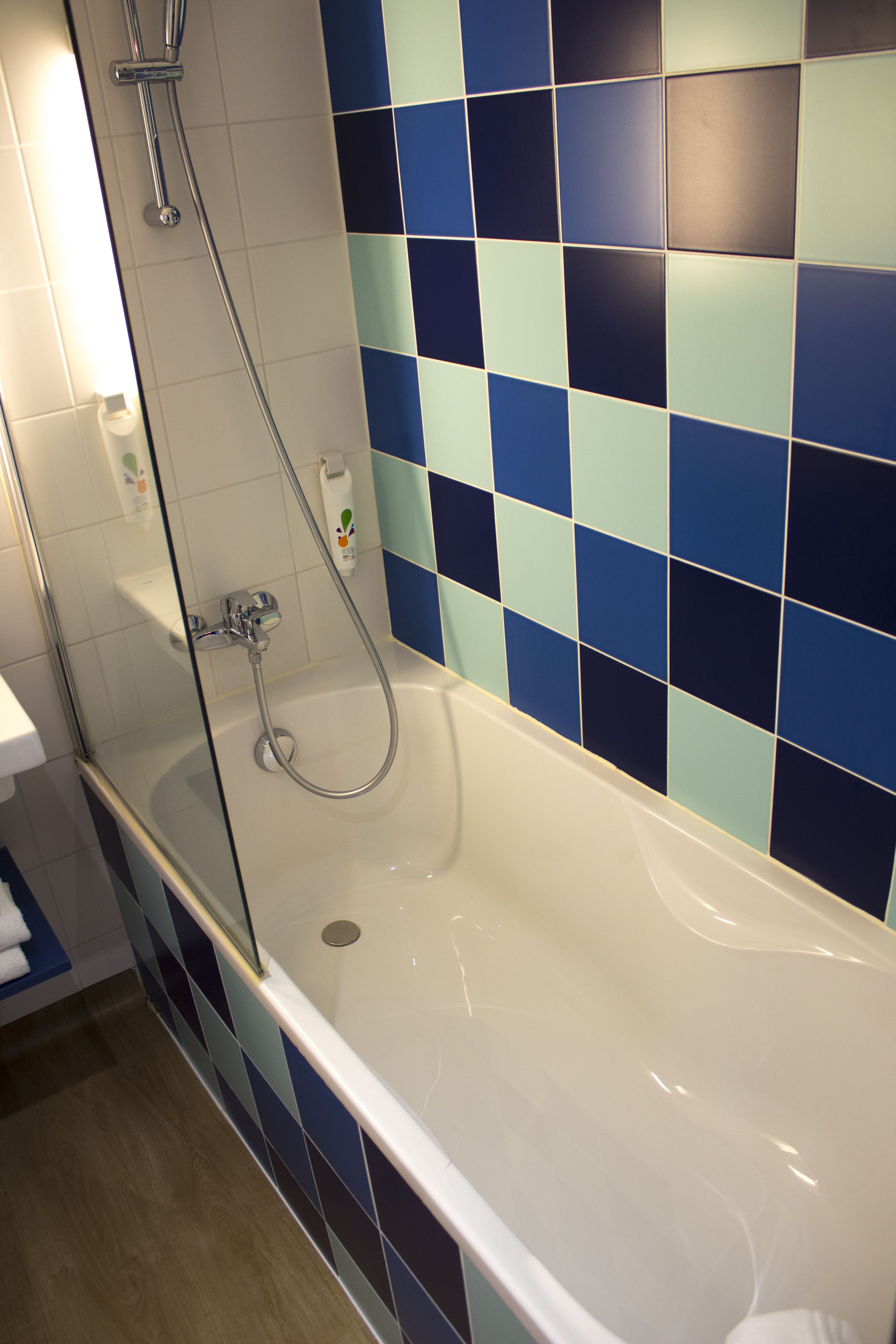 A bathroom with a glass-enclosed shower, a white bathtub, and blue and light blue tiles on the walls.