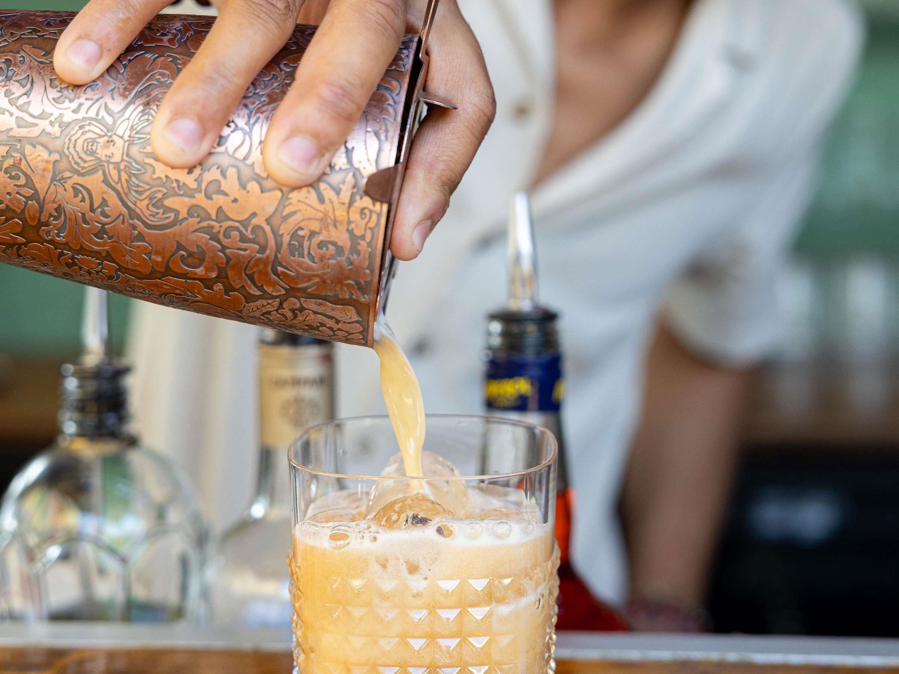 A hand pouring a drink from a copper-colored container into a glass with a textured pattern.