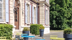 Een terras met blauwe en gele tafels en stoelen voor een gebouw met een gevel van rode baksteen en beige steen.