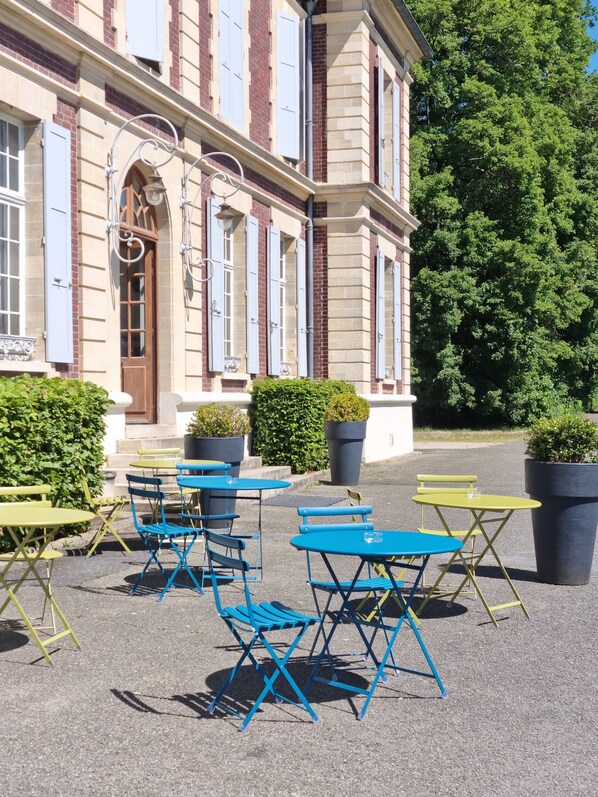 An outdoor seating area with blue and yellow tables and chairs in front of a building with red brick and beige stone facade. - Château de l'Hermitage (Ennery)
