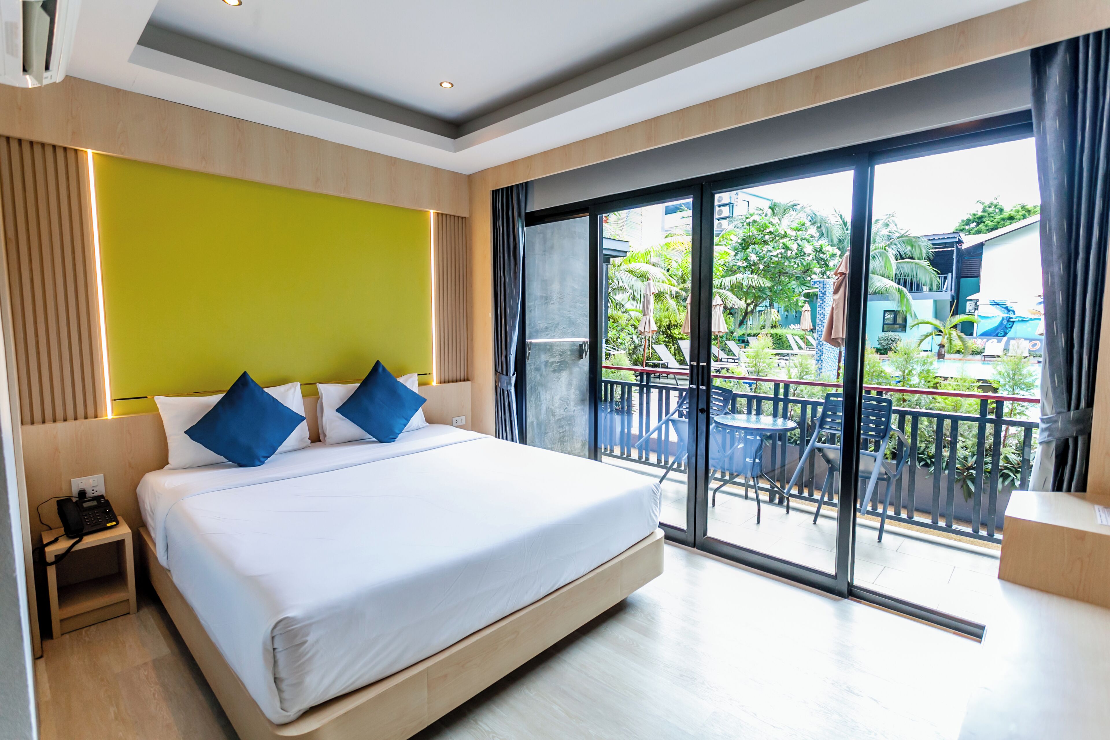A modern hotel room with a large bed, a balcony with outdoor seating, and a view of the garden and pool area.