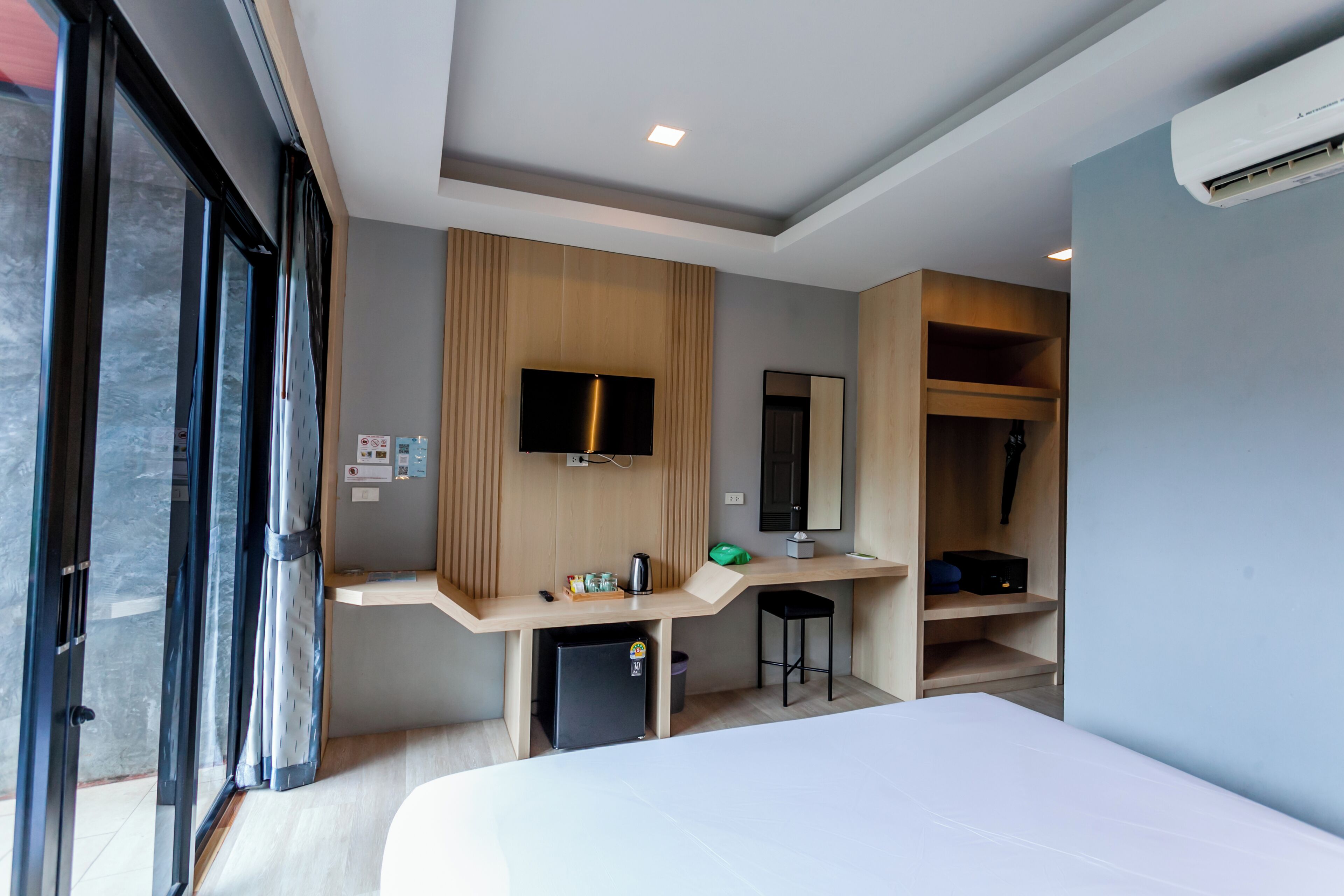 A modern hotel room with a bed, a desk with a flat-screen TV, a mini-fridge, a trash bin, and a wardrobe.