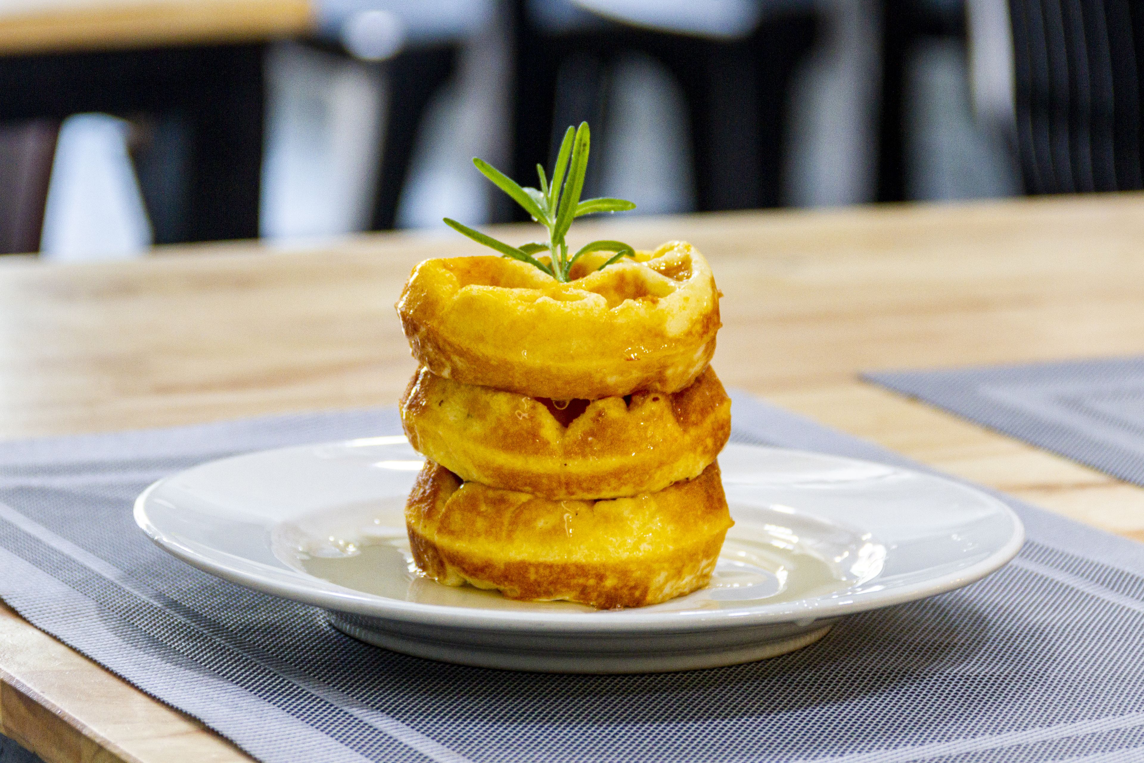 A stack of golden-brown pancakes on a white plate with a sprig of rosemary on top, placed on a table with a blue and white checkered placemat.