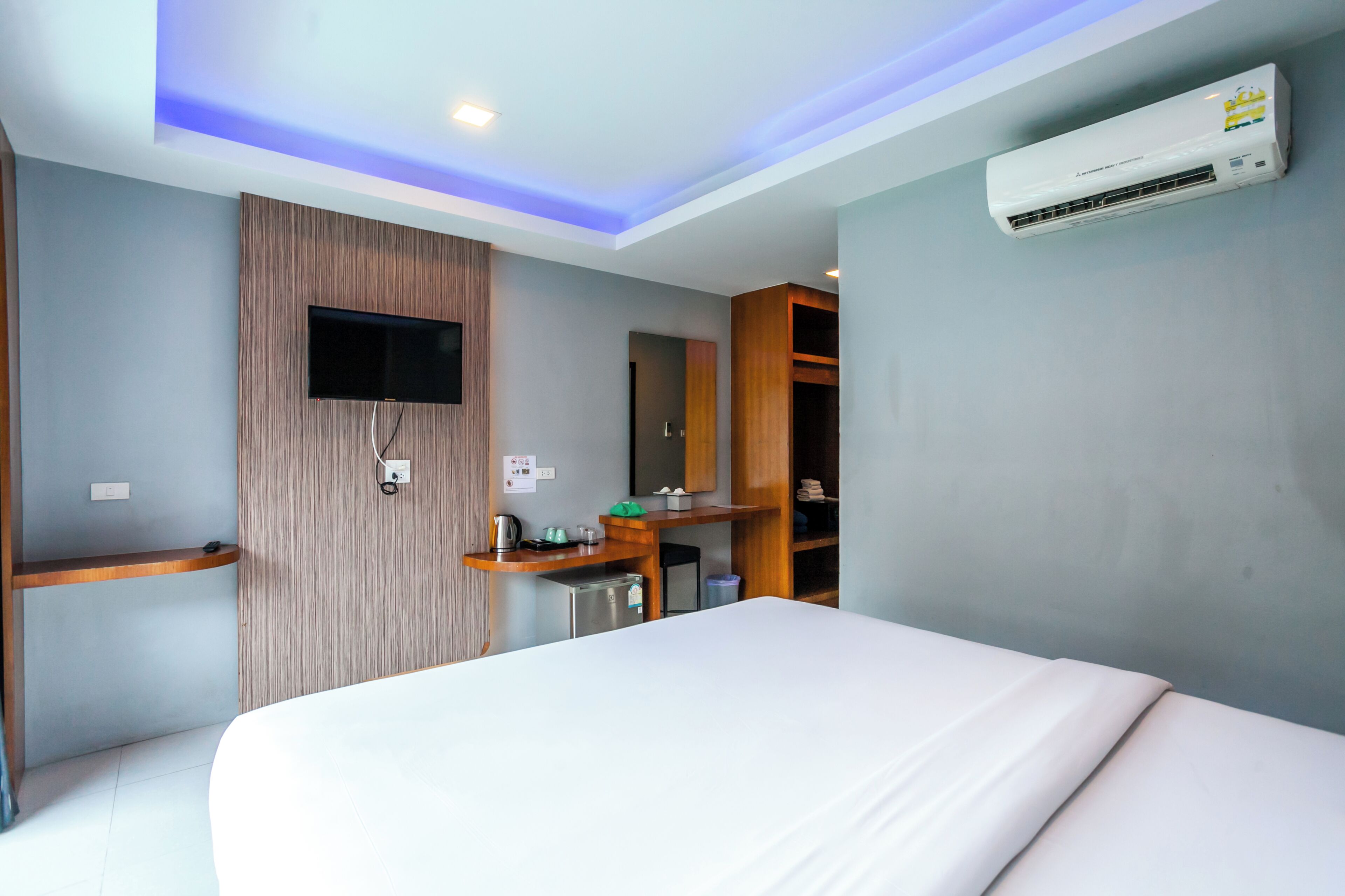 A modern hotel room with a bed, a TV mounted on the wall, a built-in desk with a kettle, and a wardrobe.