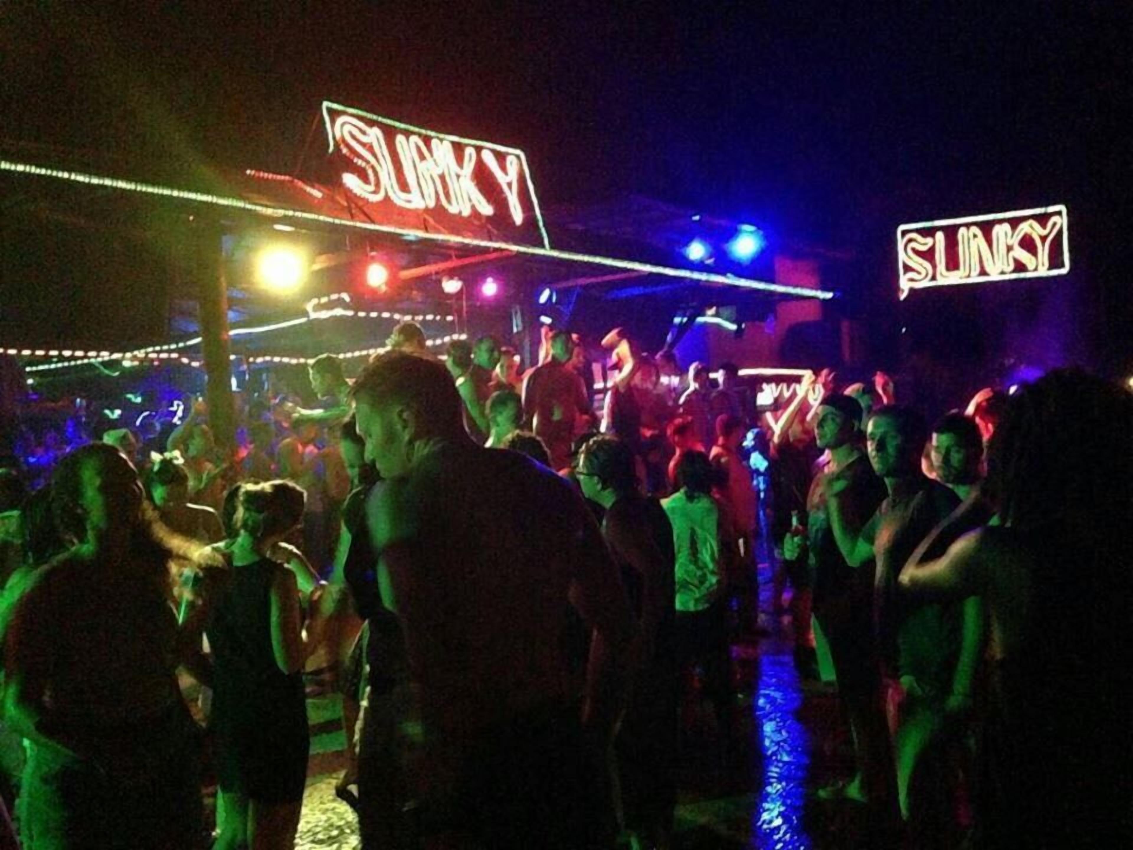 A crowded outdoor area with neon signs reading "SUNKY" and people dancing.