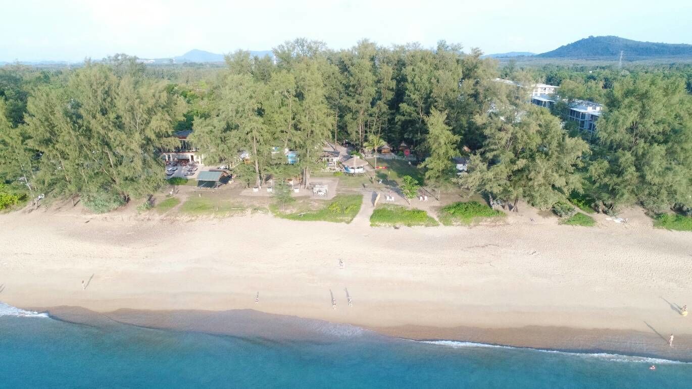 An aerial view of a beach with a dense forest in the background, a clear sky, and a few buildings near the shore.