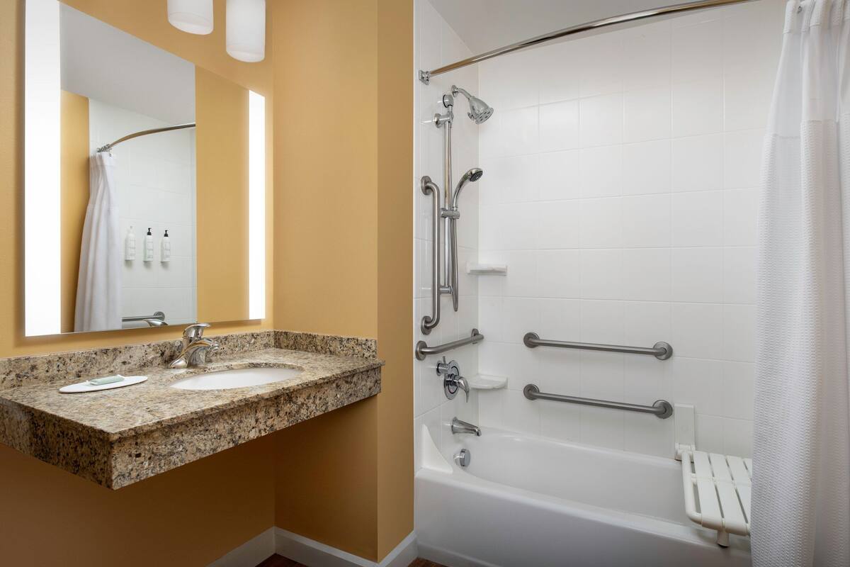 Une salle de bain avec un lavabo sur un plan de travail en granit, une baignoire blanche dotée de barres d’appui et un rideau de douche.