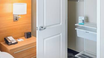 A hotel room with a bed, bedside table with a phone, a nightstand, a wall-mounted lamp, a closet with shelves, and a carpeted floor.