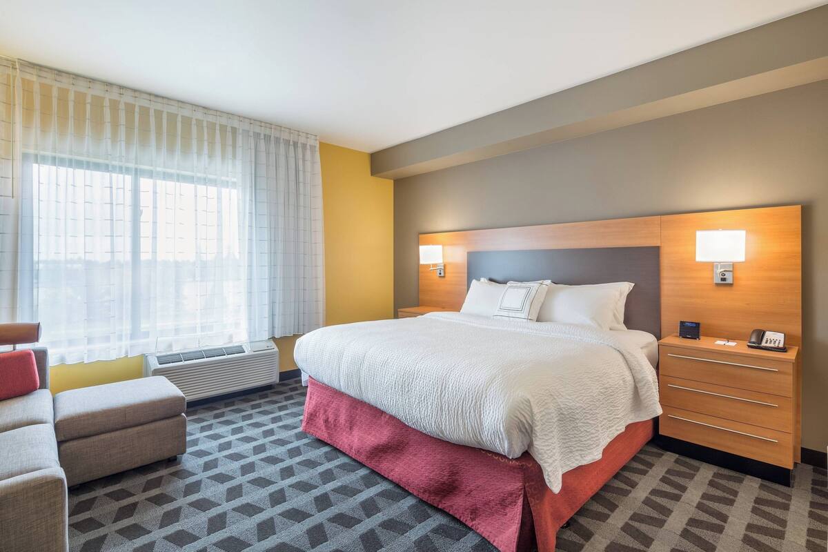 Une chambre d’hôtel avec un grand lit, une table de chevet avec une lampe, une chaise et une fenêtre avec des rideaux.