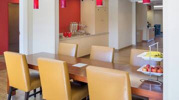 Un coin repas avec une table en bois et des chaises jaunes, un espace cuisine avec un mur rouge et des lampes suspendues rouges.