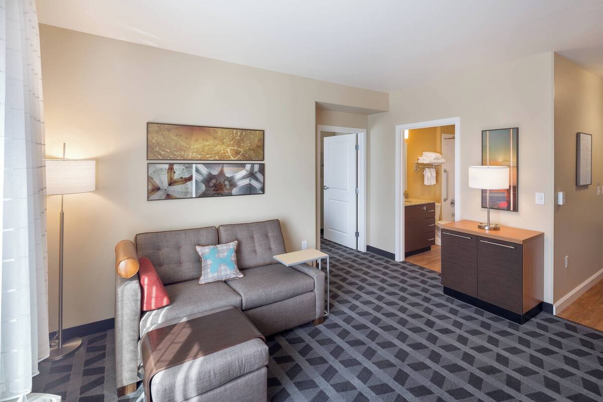Une chambre d’hôtel moderne avec un canapé gris, une petite table et un meuble. On y trouve une porte donnant accès à la salle de bain et un couloir.