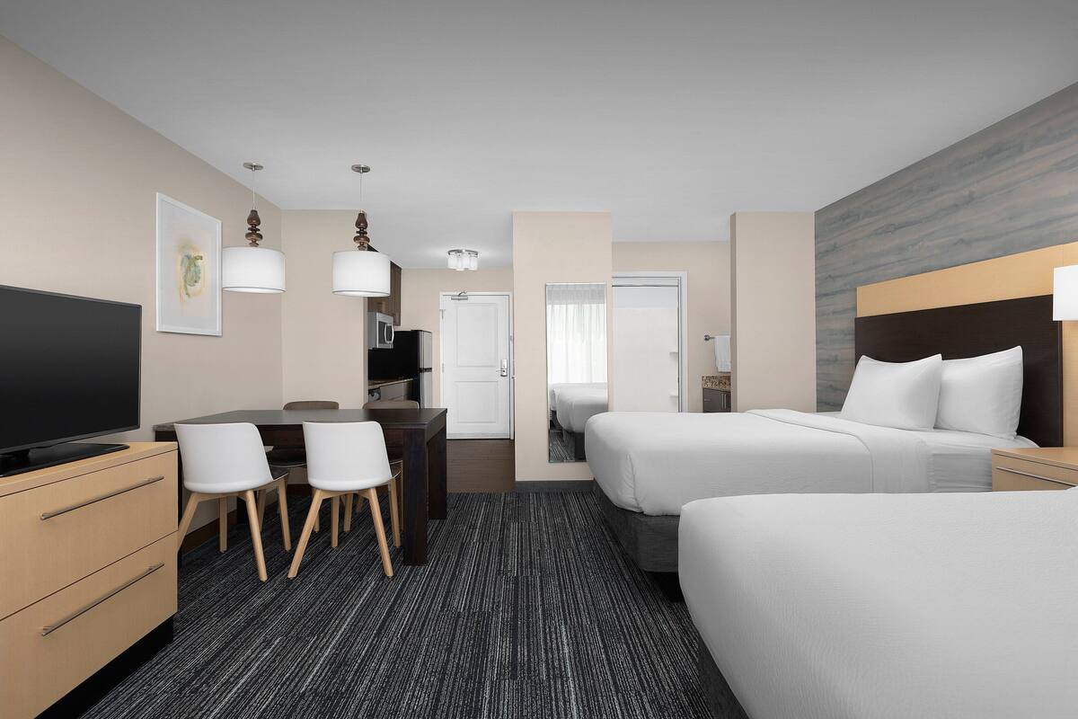 Habitación de hotel con dos camas, un escritorio con sillas, un televisor y un espejo grande.