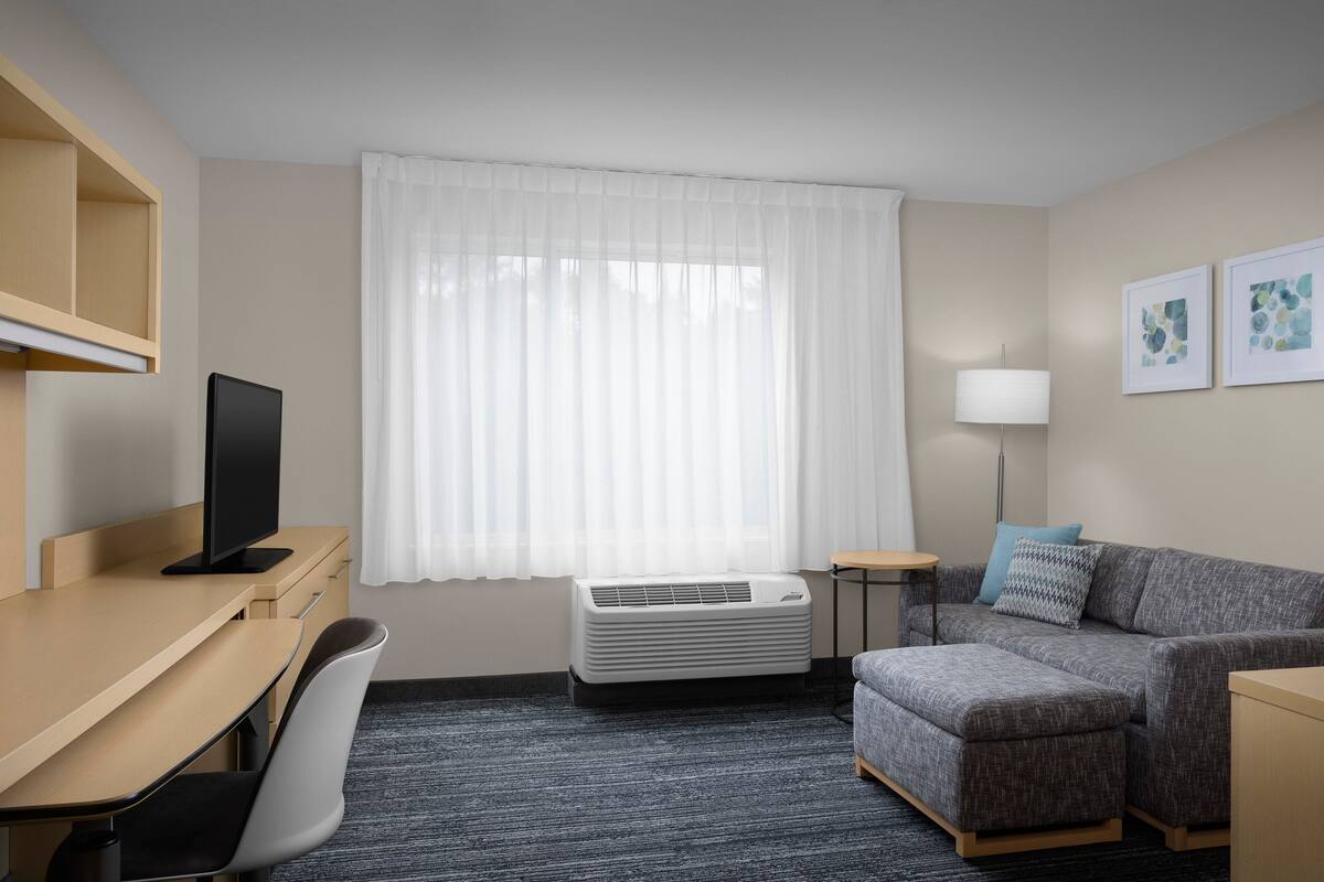 Una habitación de hotel con escritorio, televisión de pantalla plana, sofá gris, una mesita pequeña y aire acondicionado instalado en la pared.