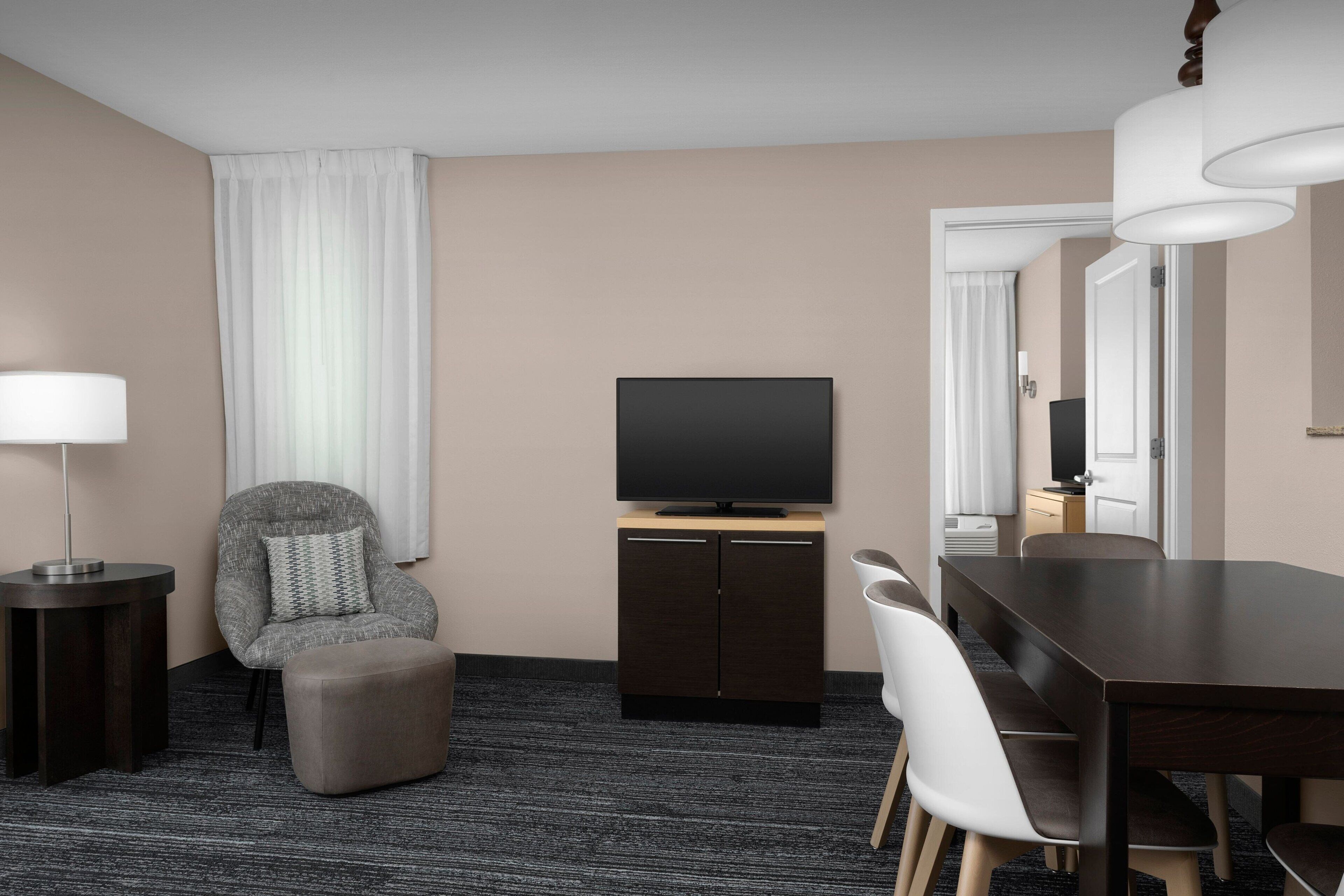 A hotel room with a dark wood dining table, a grey upholstered armchair, a flat-screen TV on a wooden cabinet, a standing lamp, and a large window with white curtains.