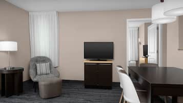 Habitación de hotel con una mesa de comedor de madera oscura, un sillón tapizado de color gris, un televisor de pantalla plana sobre un mueble de madera, una lámpara de pie y un ventanal con cortinas blancas.