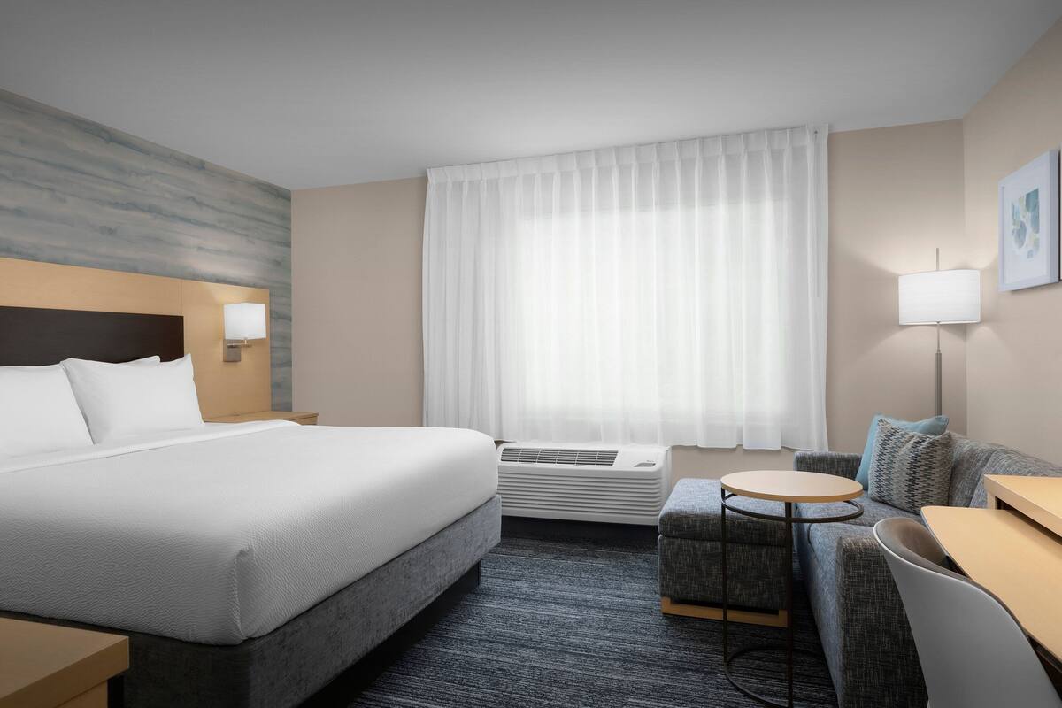 Habitación de hotel con cama, una mesita redonda, sofá, escritorio, lámpara y ventana con cortinas.