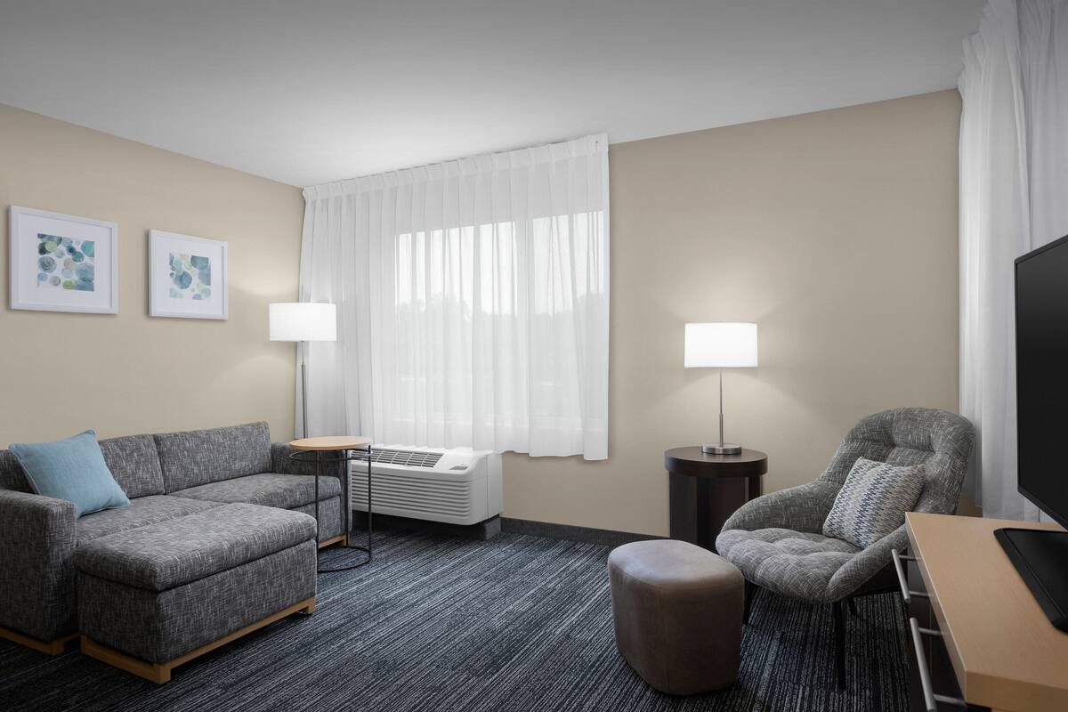 Habitación de hotel con sofá gris, sillón gris, una mesita, lámpara y televisor.