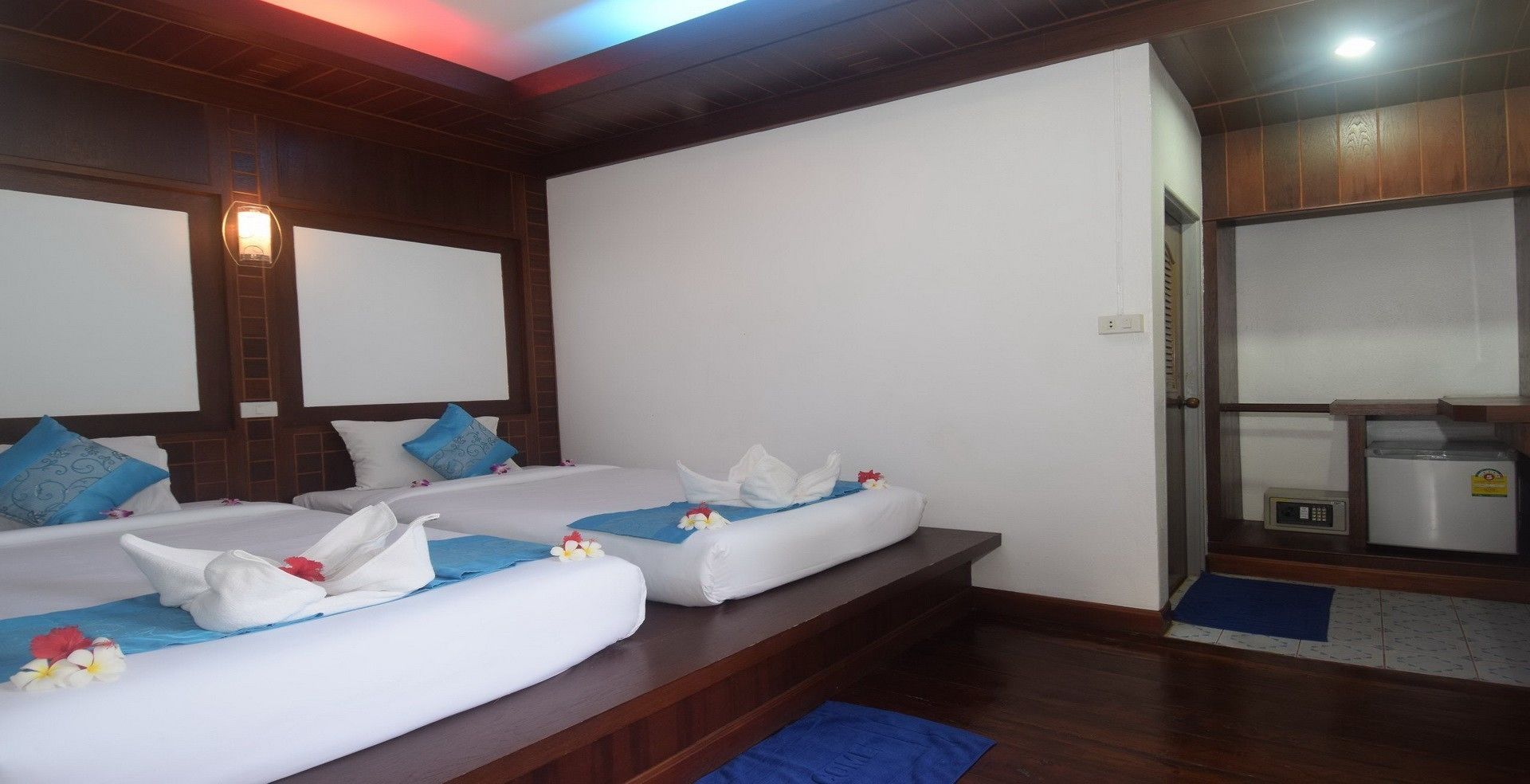 A hotel room with two beds, each with white linens and blue pillows, a small table, and a refrigerator.
