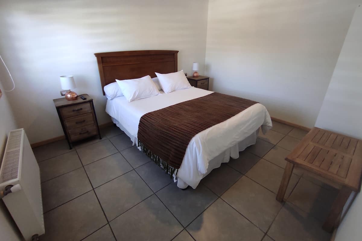 Cama bem arrumada com lençóis brancos e manta marrom, mesa de cabeceira de madeira com abajur e uma pequena mesa de madeira.